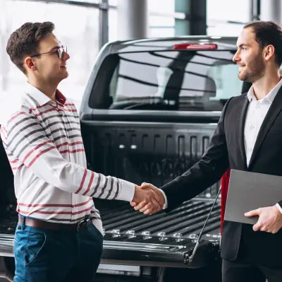 Two individuals shake hands in front of a parked pickup truck, symbolizing a business agreement or sale.