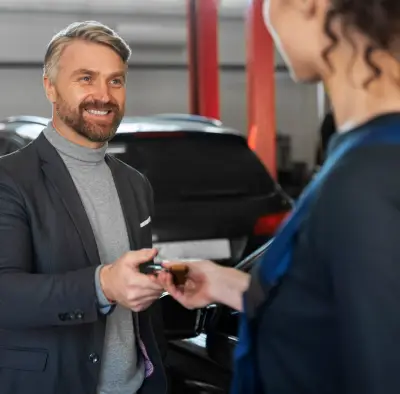 A man in a suit hands car keys to a woman in a blue dress, surrounded by parked cars in a well-lit garage.