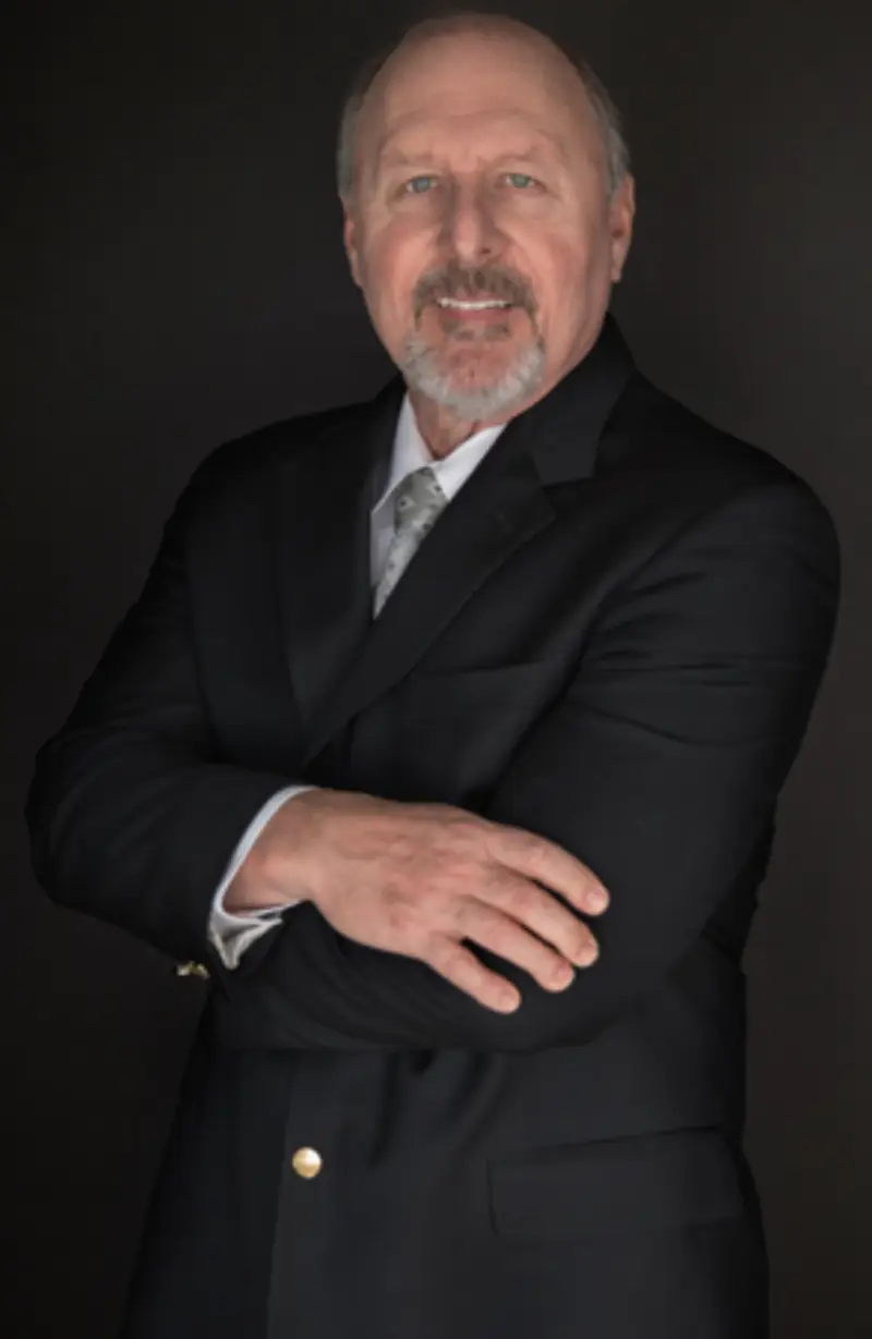 A man in a formal black suit stands confidently with his arms crossed, set against a dark background.